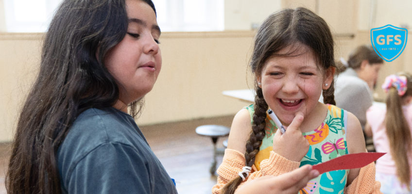 Two GFS girls - one is showing the other something she has written on a piece of paper. The other is laughing.